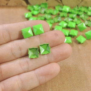 May include: A close-up of a hand holding four green metal pyramid-shaped studs. The studs are square and have a shiny finish. There are many more green studs scattered on a brown surface in the background.