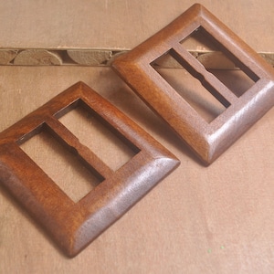 May include: Two wooden buckles with a diamond shape and a rectangular opening. The buckles are a warm brown color and appear to be made of wood. The design includes a central bar within the rectangular opening.