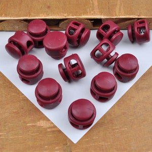 Puede incluir: Un grupo de 12 topes de cierre de plástico burdeos con un mecanismo de resorte. Los topes están dispuestos sobre una superficie blanca.