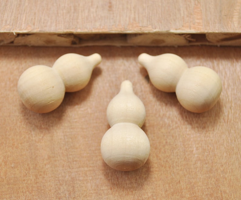 May include: Three wooden gourds, each with two bulbs, are arranged on a light brown wooden surface. The gourds are a light beige color and have a smooth, slightly textured surface.