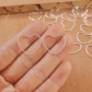 Puede incluir: Dos aros de alambre de plata en forma de corazón, sostenidos en una mano, con una pila de aros similares en el fondo.