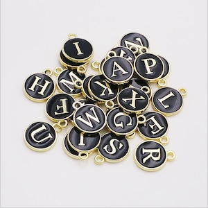 Può includere: Una collezione di ciondoli con lettere nere e dorate. Ogni ciondolo è un disco rotondo con un bordo dorato e una lettera bianca su sfondo nero.