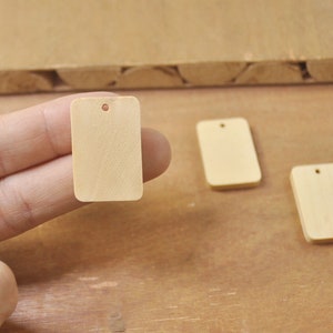 Puede incluir: Tres etiquetas rectangulares de madera sin terminar con un agujero en la parte superior. Las etiquetas son de color marrón claro y están sobre una superficie de madera.