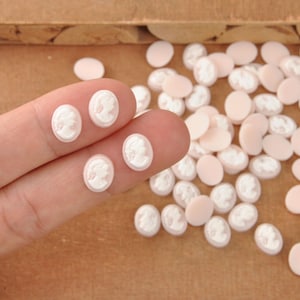 May include: Oval-shaped cameo cabochons in a light pink hue with a white profile design. Several are scattered on a wooden surface, with four held between fingers. These craft supplies are ideal for jewelry making or embellishments.