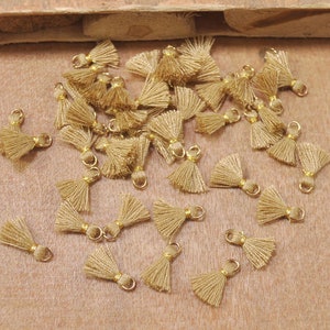 May include: A collection of small, gold-colored tassels with a loop at the top. The tassels are made of a soft, beige fabric and are arranged in a random pattern on a brown surface.