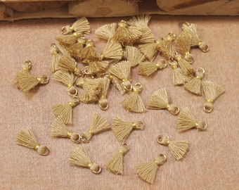May include: A collection of small, gold-colored tassels with a loop at the top. The tassels are made of a soft, beige fabric and are arranged in a random pattern on a brown surface.