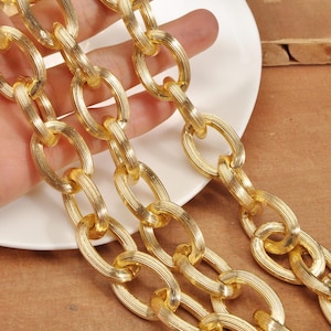 May include: Close-up of a gold-colored chain with large, oval links. The chain has a textured surface, and the links are interlocked. The chain is displayed on a white plate and a brown surface.