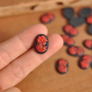 May include: Oval-shaped cameo cabochons with a black background and a red profile of a woman. The cabochons are small, with a smooth surface, and are scattered across a wooden surface. The cameo design is detailed and classic.