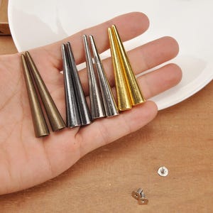 Op de afbeelding: Een verzameling metalen conische studs in brons, zilver en goudtinten. De studs worden in een hand gehouden, met een wit bord op de achtergrond. Kleine zilveren schroeven zijn ook zichtbaar.
