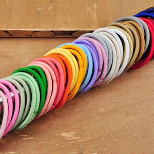 K&ouml;nnte beinhalten: Ein gro&szlig;er Stapel bunter Haargummis in verschiedenen Farbt&ouml;nen von Rosa, Gr&uuml;n, Gelb, Blau, Lila, Braun und Rot. Die Haargummis sind spiralf&ouml;rmig angeordnet, wobei die dunkelsten Farben unten und die hellsten Farben oben liegen.