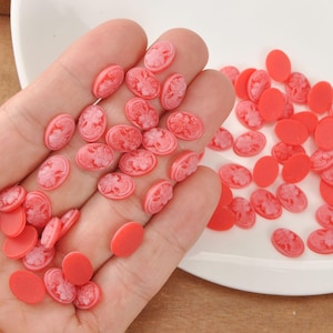 May include: A collection of small, oval-shaped, red and white cameo beads. The beads feature a profile of a woman and are scattered across a white surface. Some beads are solid red, while others have a white cameo design.