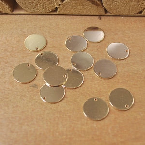 May include: A collection of small, gold-colored, round metal charms with a hole at the top. The charms are scattered on a light brown wooden surface. These charms are ideal for jewelry making or craft projects.