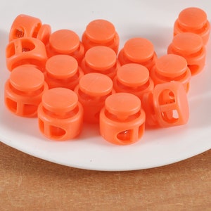 May include: A group of 15 orange plastic toggle clasps, also known as cord locks, are arranged in a loose pile on a white plate. The clasps have a round shape with a hole in the centre and two holes on the sides.