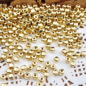 May include: A large quantity of gold-coloured square beads. The beads have a hole in the centre and are scattered on a white lace surface.