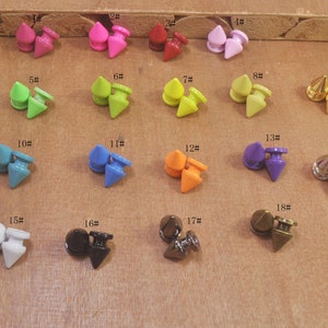 May include: Assortment of 18 colourful and metallic pyramid-shaped studs with a pointed tip. The studs are arranged in rows on a wooden surface. Each stud has a number next to it, ranging from 1 to 18.