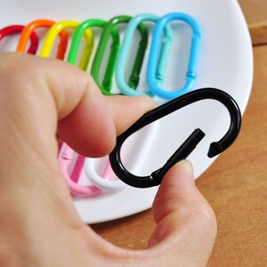May include: A black metal key chain with a spring-loaded clasp. The keychain is being held by a hand. The keychain is surrounded by a variety of colorful keychains.