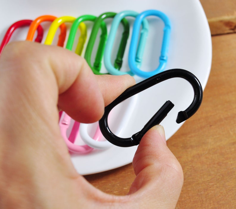 May include: A black metal key chain with a spring-loaded clasp. The keychain is being held by a hand. The keychain is surrounded by a variety of colorful keychains.