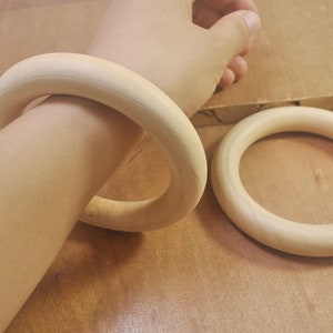 May include: Two wooden rings, one worn on a wrist and the other lying on a wooden surface. Both rings are made of light-colored wood and have a smooth, polished finish.