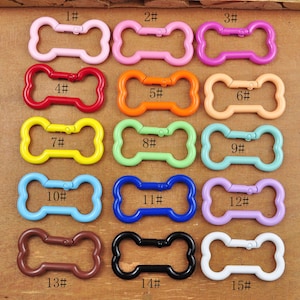 Op de afbeelding: Vijftien kleurrijke, botvormige metalen sluitingen met de nummers 1 tot en met 15. De sluitingen zijn in verschillende kleuren, waaronder roze, oranje, rood, geel, groen, blauw, paars, bruin, zwart en wit.
