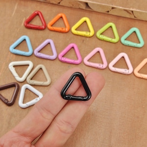 Può includere: Una collezione di anelli metallici triangolari colorati in varie tonalità, tra cui rosso, arancione, giallo, verde, blu, viola, rosa, bianco, marrone e nero. Gli anelli sono disposti su una superficie di legno.