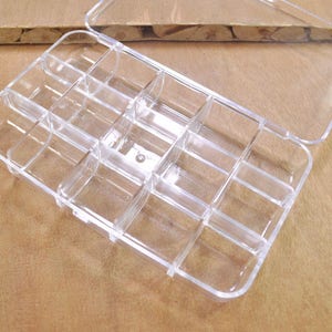 May include: A clear plastic storage box with a hinged lid and 12 individual compartments. The box is empty and ready to be filled with small items.