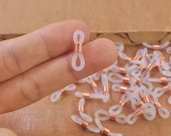 Puede incluir: Un conector de cadena de gafas de plástico blanco con un acento de metal dorado rosa. El conector se sostiene en una mano, y se puede ver una pila de conectores similares en el fondo.