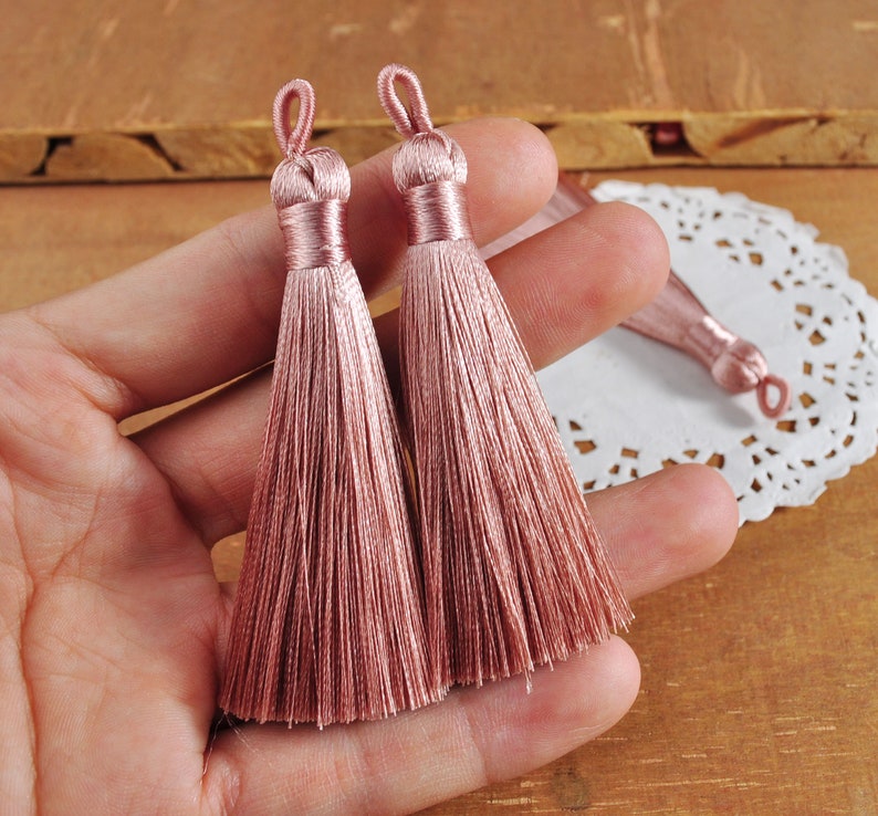 May include: Two dusty rose silk tassels with a loop at the top. The tassels are made of soft, fine threads and are approximately 2 inches long.