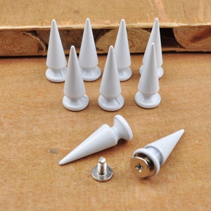 Puede incluir: Diez tachuelas de metal blanco en forma de pirámide con punta puntiaguda. Las tachuelas tienen una base plana y están dispuestas en círculo. Se muestran dos tachuelas adicionales por separado, una con un tornillo y otra sin.