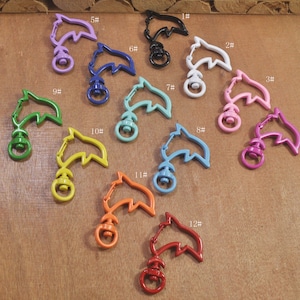 Puede incluir: Doce llaveros con forma de delfín en varios colores, incluyendo morado, azul, blanco, rosa, verde, amarillo, naranja y rojo. Cada llavero tiene un anillo de metal unido a la cola del delfín.
