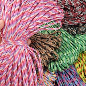 May include: A hand holds a bundle of pink, red, and white striped paracord, tied with a matching cord. Other bundles of paracord in various colors, including green, brown, and red, are visible in the background. The cords are tightly wound.
