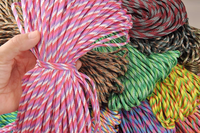 May include: A hand holds a bundle of pink, red, and white striped paracord, tied with a matching cord. Other bundles of paracord in various colors, including green, brown, and red, are visible in the background. The cords are tightly wound.