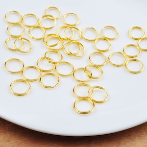 May include: A collection of gold jump rings, a type of jewellery finding, scattered on a white surface.