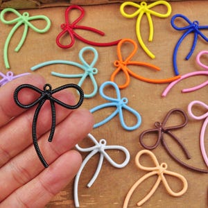 Peut inclure: Assortiment de breloques en métal en forme de nœud, de différentes couleurs, dont le rouge, le bleu, le vert, le jaune, le rose et le noir. Les breloques sont en forme de nœuds avec de longues queues. Les breloques sont présentées sur une surface en bois.