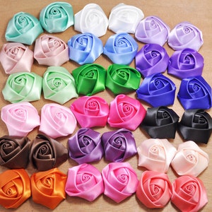 May include: Assortment of 24 satin ribbon roses in various colors, including pink, blue, green, purple, orange, brown, black, and white. The roses are arranged in a square pattern on a wooden surface.