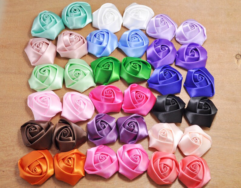 May include: Assortment of 24 satin ribbon roses in various colors, including pink, blue, green, purple, orange, brown, black, and white. The roses are arranged in a square pattern on a wooden surface.