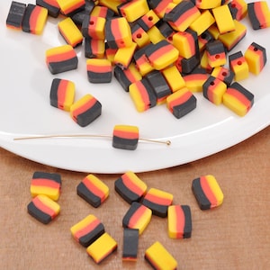Könnte beinhalten: Eine Ansammlung kleiner, rechteckiger Perlen in Gelb, Rot und Schwarz, die an die deutsche Flagge erinnern. Die Perlen liegen auf einem weißen Teller und einer Holzoberfläche. Ein goldfarbener Draht verläuft durch eine Perle.
