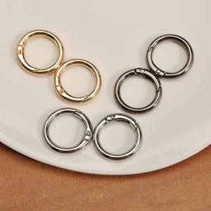 May include: A collection of round metal spring snap rings in gold and silver tones. The rings are open, showing the spring mechanism. The snap rings are on a white surface, suitable for crafting or jewelry making.