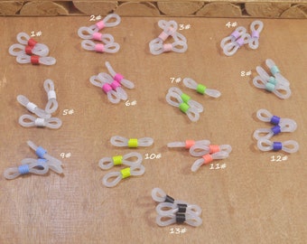 Puede incluir: Una colección de retenedores de gafas claros y flexibles con acentos coloridos. Cada retenedor presenta un diseño de bucle con una banda de color en varios tonos, incluyendo rojo, rosa, verde y azul. Los retenedores se muestran sobre una superficie marrón claro.