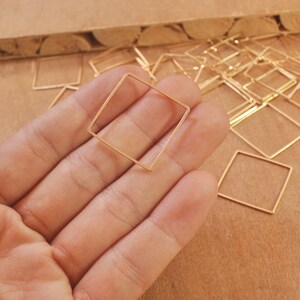Puede incluir: Primer plano de una mano sosteniendo un marco cuadrado de alambre dorado. El marco mide aproximadamente 2,5 cm de lado y está hecho de alambre fino. Hay muchos otros marcos similares en el fondo.