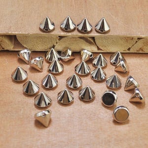 May include: A selection of silver-coloured, cone-shaped metal studs with a pointed top and a flat base. The studs are arranged on a light brown surface, with some showing a hole for attachment. Ideal for craft projects.