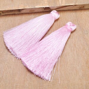 May include: Two pink silk tassels with a long fringe. The tassels are on a wooden surface.
