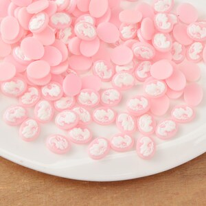 May include: A collection of pink and white cameo cabochons. The cabochons are oval-shaped with a raised white cameo design on a pink background. The cabochons are scattered on a white plate, creating a delicate and feminine aesthetic.