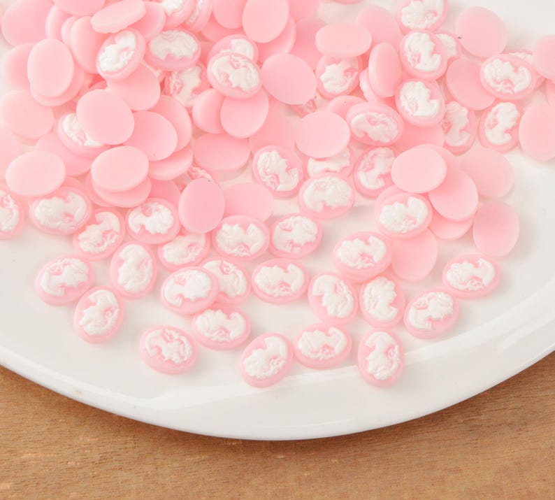 May include: A collection of pink and white cameo cabochons. The cabochons are oval-shaped with a raised white cameo design on a pink background. The cabochons are scattered on a white plate, creating a delicate and feminine aesthetic.
