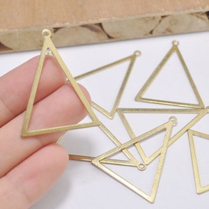 May include: Several brass-colored, open-frame triangle charms with small loops at the top for jewelry making. The charms are displayed on a white surface, with one held in a hand. The triangles are approximately 1 inch in height.