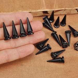 May include: A selection of black, conical body jewellery, some with a flared base, displayed on a wooden surface. Several pieces are held in a hand, with others scattered around. Small silver screws are also present.