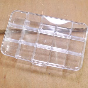 May include: A clear plastic storage box with 12 compartments. The box is empty and has a rectangular shape.