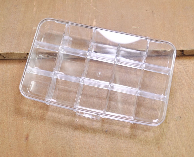 May include: A clear plastic storage box with 12 compartments. The box is empty and has a rectangular shape.