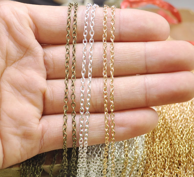Puede incluir: Primer plano de cinco estilos de cadena diferentes en varios acabados met&aacute;licos, incluyendo bronce, plata y oro. Las cadenas se sostienen en una mano, mostrando su longitud y detalle.