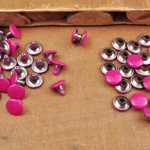 May include: A collection of pink metal rivets with silver accents. The rivets are arranged in two groups, one with the flat side facing up and the other with the pointed side facing up.