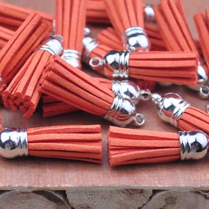 May include: A group of orange suede tassels with silver metal caps. The tassels are arranged in a pile on a wooden surface.